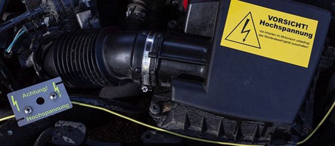 Der Motorraum eines Autos mit einem gelben Aufkleber, der vor elektrischen Gefahren warnt, und einer kleinen Karte mit der Aufschrift "Hochspannung".