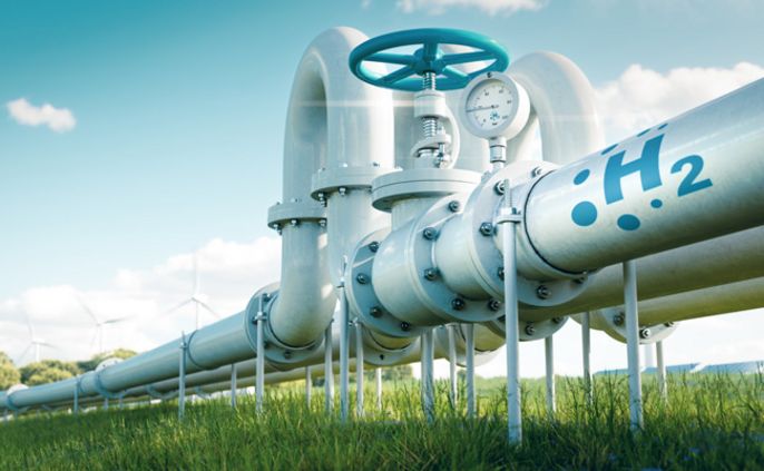 Ein großes Rohrsystem mit der Aufschrift "H2" und einem Messgerät auf einem offenen Feld mit Windkraftanlagen im Hintergrund.