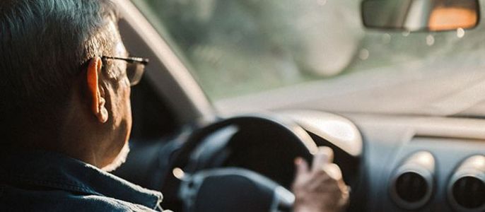 Ein älterer Mann am Steuer eines Autos, mit dem Rücken zur Kamera, trägt eine Brille und ein blaues Hemd.