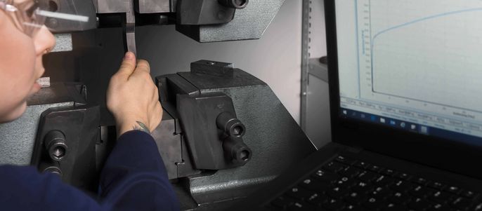 Eine Person mit Schutzbrille, die eine Maschine einstellt, während im Hintergrund eine Grafik auf einem Laptop-Bildschirm zu sehen ist.