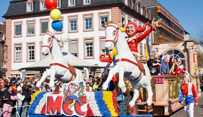 Der Mainzer Rosenmontagsumzug mit einem großen Wagen mit zwei weißen Pferden, einem Clown und verschiedenen bunten Elementen vor der Kulisse historischer Gebäude.