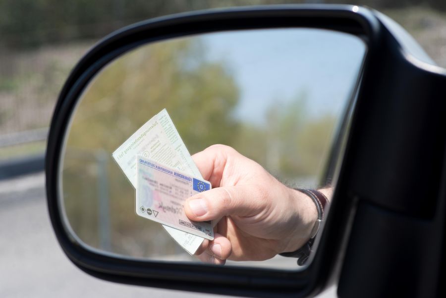 Eine Hand, die einen Führerschein hält, wobei sich der Führerschein im Seitenspiegel des Autos spiegelt.