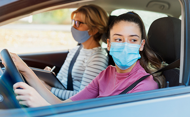 Zwei Frauen in einem Auto, beide mit Gesichtsmasken, wobei die Frau vorne das Lenkrad hält.