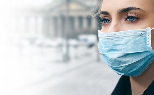 Nahaufnahme eines Frauengesichts mit einer blauen chirurgischen Maske vor einem verschwommenen Hintergrund einer Stadtlandschaft.