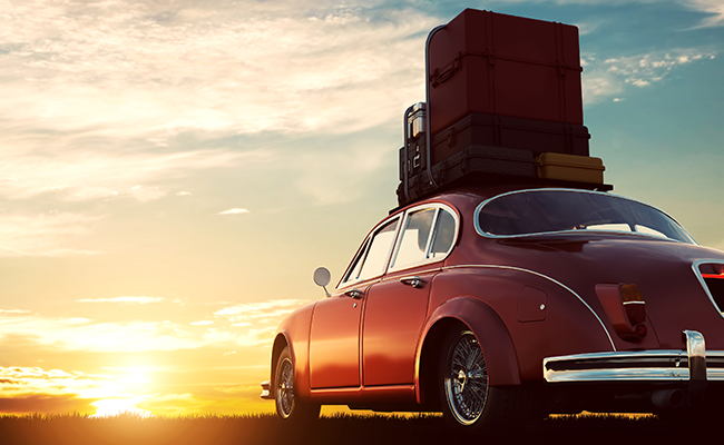 Ein roter Oldtimer, der bei Sonnenuntergang auf einem freien Feld geparkt ist und auf dessen Dach ein Stapel Koffer liegt.