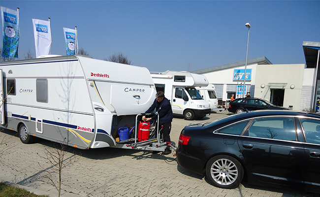 Ein weißer Wohnwagen. Ein Mann ist in der Nähe des Wohnwagens zu sehen, und im Hintergrund sind weitere Fahrzeuge und ein Geschäft zu sehen.