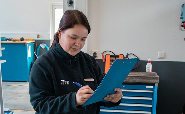 Eine Frau in einer Werkstatt, die eine schwarze Jacke mit einem Logo trägt und auf einem blauen Klemmbrett schreibt.